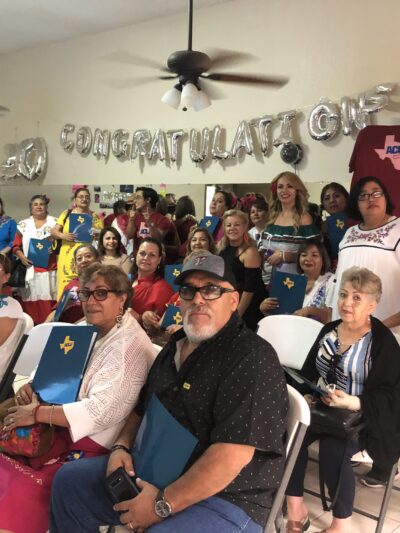 Image: A large group of people of various ages are assembled. Some sit in folding chairs, and some stand, towards the back against a wall. Many are holding folders with the ACLU of Texas logo on it. They gaze towards the camera.