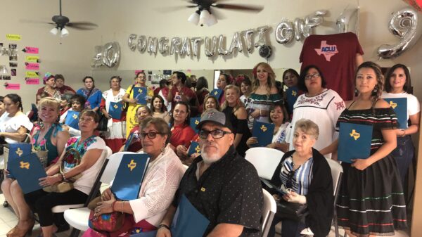 Image: A large group of people of various ages are assembled. Some sit in folding chairs, and some stand, towards the back against a wall. Many are holding folders with the ACLU of Texas logo on it. They gaze towards the camera.