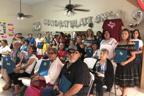 Image: A large group of people of various ages are assembled. Some sit in folding chairs, and some stand, towards the back against a wall. Many are holding folders with the ACLU of Texas logo on it. They gaze towards the camera.