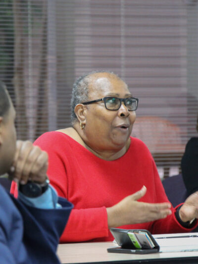 Image: People sit behind desks at different angles. In the foreground is an African-American man with his back towards the camera He looks towards an African-American woman centered in the photo. She gestures and speaks to someone not pictured.