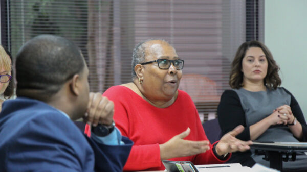 Image: People sit behind desks at different angles. In the foreground is an African-American man with his back towards the camera He looks towards an African-American woman centered in the photo. She gestures and speaks to someone not pictured.