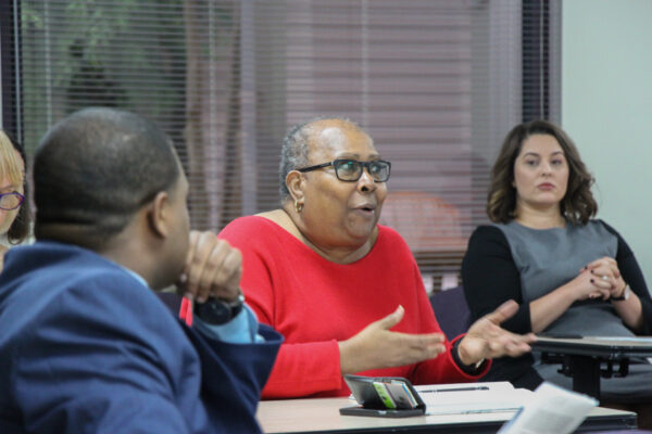 Image: People sit behind desks at different angles. In the foreground is an African-American man with his back towards the camera He looks towards an African-American woman centered in the photo. She gestures and speaks to someone not pictured.