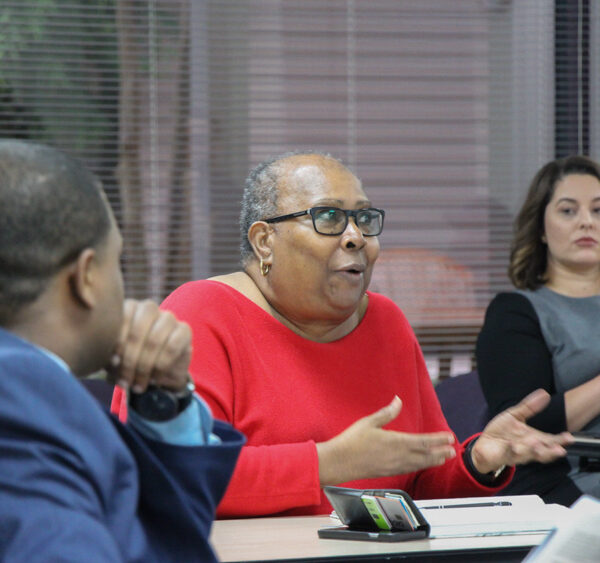 Image: People sit behind desks at different angles. In the foreground is an African-American man with his back towards the camera He looks towards an African-American woman centered in the photo. She gestures and speaks to someone not pictured.