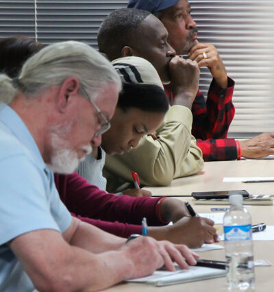 Image: A row of people of varying ages sit behind a long desk. Some have their heads down and write on paper, some face forward and look contemplative.