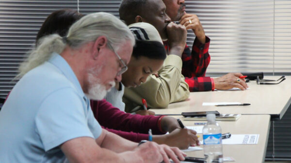 Image: A row of people of varying ages sit behind a long desk. Some have their heads down and write on paper, some face forward and look contemplative.