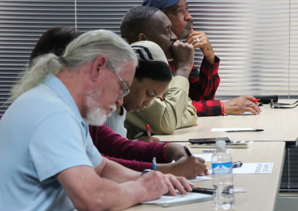 Image: A row of people of varying ages sit behind a long desk. Some have their heads down and write on paper, some face forward and look contemplative.
