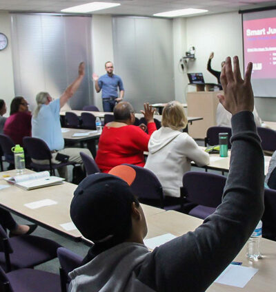 Image: Peope sit in rows behind desks, their backs facing the camera. Several raise their hands and look forward to the front of a room. A powerpoint is projected onto a pull-down screen, A man standing up faces them.