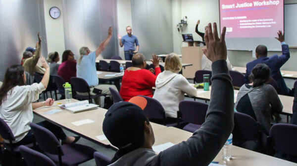 Image: Peope sit in rows behind desks, their backs facing the camera. Several raise their hands and look forward to the front of a room. A powerpoint is projected onto a pull-down screen, A man standing up faces them.