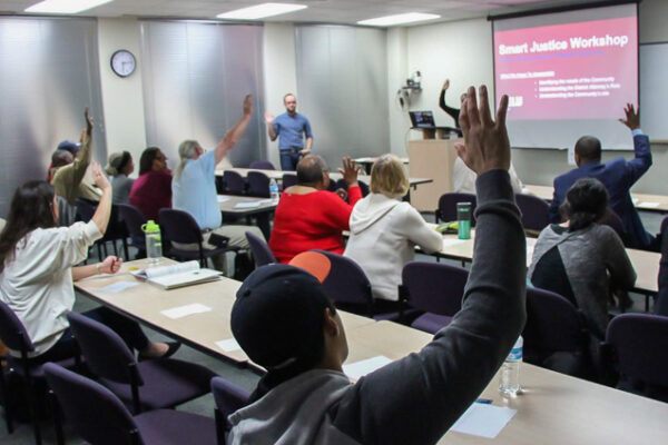Image: Peope sit in rows behind desks, their backs facing the camera. Several raise their hands and look forward to the front of a room. A powerpoint is projected onto a pull-down screen, A man standing up faces them.