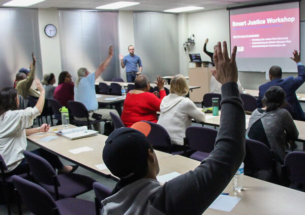 Image: Peope sit in rows behind desks, their backs facing the camera. Several raise their hands and look forward to the front of a room. A powerpoint is projected onto a pull-down screen, A man standing up faces them.