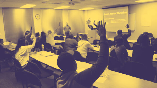 Image: People sit in two sets of rows behind long desks. Their backs face the camera. Several are raising their hands and looking across a room at a someone standing up and addressing them.