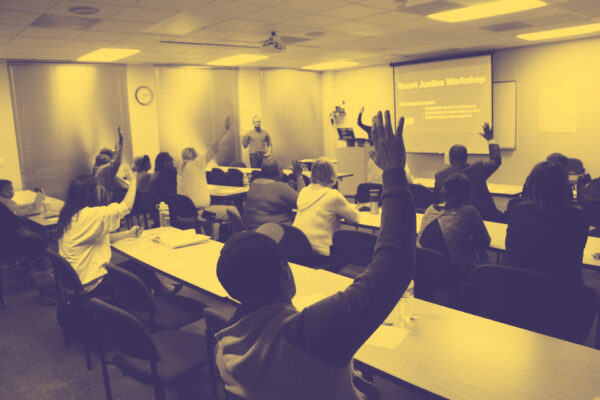 Image: People sit in two sets of rows behind long desks. Their backs face the camera. Several are raising their hands and looking across a room at a someone standing up and addressing them.