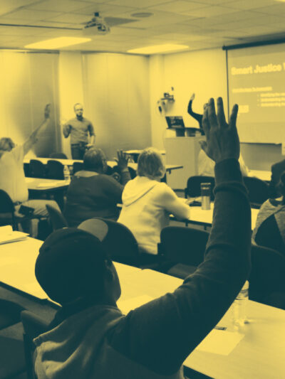 Image: People sit in two sets of rows behind long desks. Their backs face the camera. Several are raising their hands and looking across a room at a someone standing up and addressing them.