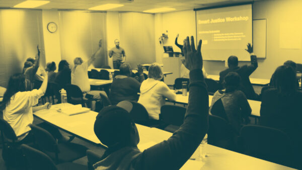 Image: People sit in two sets of rows behind long desks. Their backs face the camera. Several are raising their hands and looking across a room at a someone standing up and addressing them.