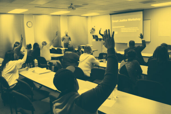 Image: People sit in two sets of rows behind long desks. Their backs face the camera. Several are raising their hands and looking across a room at a someone standing up and addressing them.