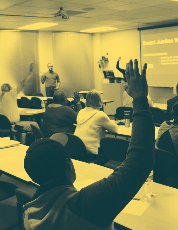 Image: People sit in two sets of rows behind long desks. Their backs face the camera. Several are raising their hands and looking across a room at a someone standing up and addressing them.