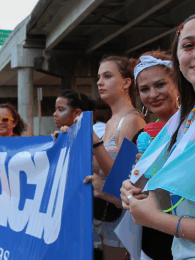 Image: Several people are dressed festively. They stand next to one another under an overpass. Some hold up an ACLU of Texas banner.One has glitter and face paint on and a transgender pride flag draped round her shoulders. Two of them pose for the camera.