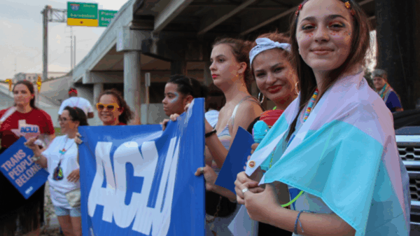 Image: Several people are dressed festively. They stand next to one another under an overpass. Some hold up an ACLU of Texas banner.One has glitter and face paint on and a transgender pride flag draped round her shoulders. Two of them pose for the camera.