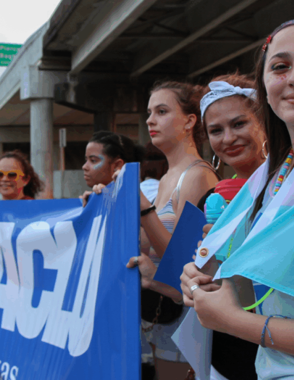 Image: Several people are dressed festively. They stand next to one another under an overpass. Some hold up an ACLU of Texas banner.One has glitter and face paint on and a transgender pride flag draped round her shoulders. Two of them pose for the camera.