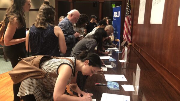 Image: A large group congregates by a long desk. Some wait as others lean down and write on postcards.