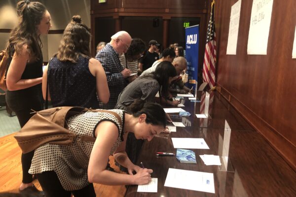 Image: A large group congregates by a long desk. Some wait as others lean down and write on postcards.