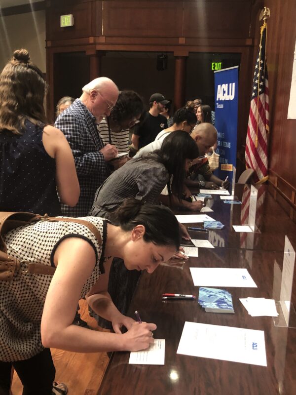 Image: A large group congregates by a long desk. Some wait as others lean down and write on postcards.