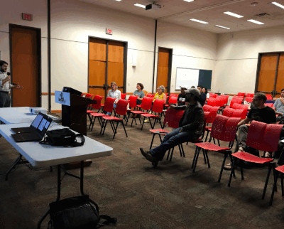 Image: Several people are spread out over rows of chairs. They face in one direction, looking at a man with a microphone. He addresses them and stand next to a podium, folding table and laptop.