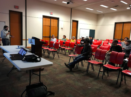 Image: Several people are spread out over rows of chairs. They face in one direction, looking at a man with a microphone. He addresses them and stand next to a podium, folding table and laptop.