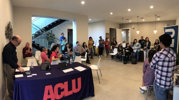Image: A photo shows an open room. Many people sit in chairs, stand or lean on tables as they look to an unpictured person speaking. Closer to the camera is a table with an ACLU of Texas tablecloth draped over it with handouts on the table.