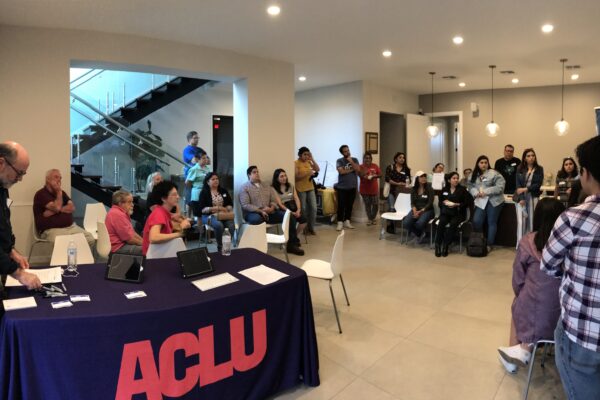 Image: A photo shows an open room. Many people sit in chairs, stand or lean on tables as they look to an unpictured person speaking. Closer to the camera is a table with an ACLU of Texas tablecloth draped over it with handouts on the table.