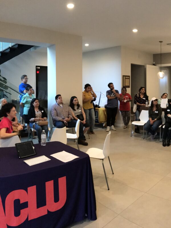 Image: A photo shows an open room. Many people sit in chairs, stand or lean on tables as they look to an unpictured person speaking. Closer to the camera is a table with an ACLU of Texas tablecloth draped over it with handouts on the table.