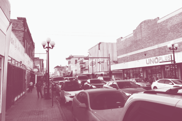 Image: A stylized photo of downtown Brownsville with cars parked one after the other on both sides of the street curb. Low-rise buildings stand on both sides.