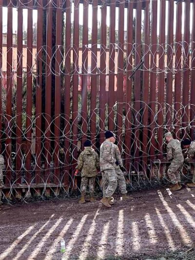 US military members are seen next to a red metal and barbed wire fence at the US border
