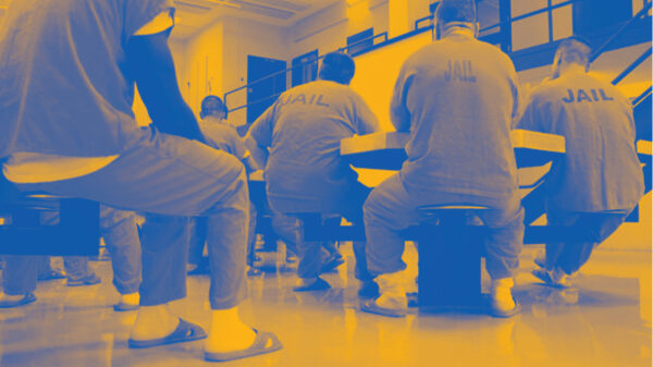 Image: A stylized photo shows men wearing uniforms that say "Jail" on their backs. They sit around tables in a carceral facility