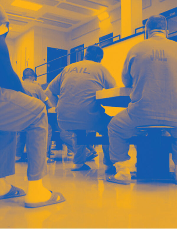 Image: A stylized photo shows men wearing uniforms that say "Jail" on their backs. They sit around tables in a carceral facility