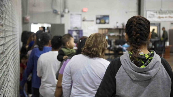 Image: A photograph shows people queued up in a large detention facility. Their backs face the camera.