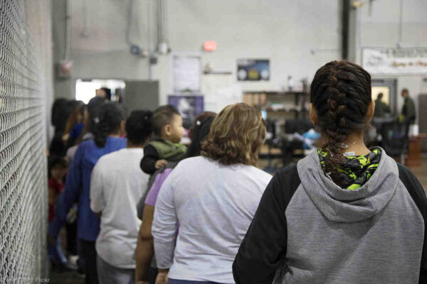 Image: A photograph shows people queued up in a large detention facility. Their backs face the camera.