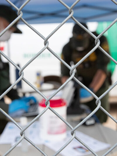 CBP agents wearing PPE processing someone wearing a mask behind a chain-link fence.