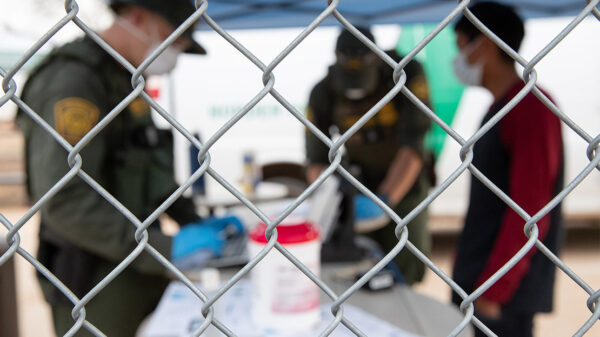 CBP agents wearing PPE processing someone wearing a mask behind a chain-link fence.