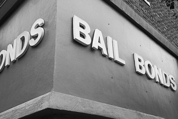 Image: A black and white photo shows the sign on a storefront located on the corner of a building. The sign extends across two sides of the building, on both sides, it advertises, "Bail Bonds."