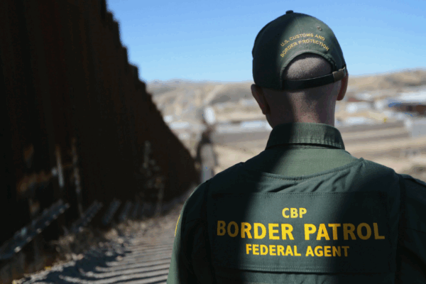 A photo shows a Border Patrol officer standing in the shadow of a border barrier. His back faces the camera and the barrier extends all the way to the horizon.