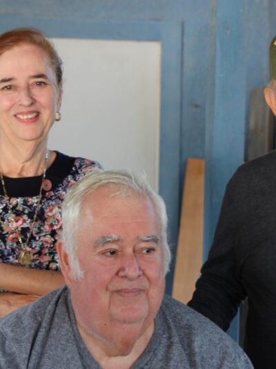 Image: A group of three pose for a photo outside of a wooden house, two standing and one sitting. The woman leans on the chair one man is sitting on. The man standing wears a baseball cap indicating he is a Vietnam War vet.