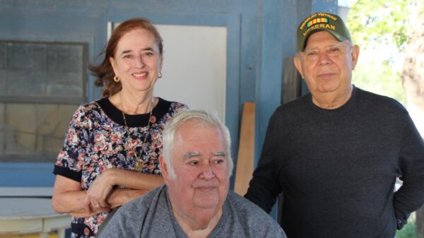 Image: A group of three pose for a photo outside of a wooden house, two standing and one sitting. The woman leans on the chair one man is sitting on. The man standing wears a baseball cap indicating he is a Vietnam War vet.