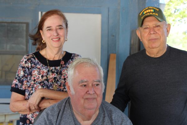 Image: A group of three pose for a photo outside of a wooden house, two standing and one sitting. The woman leans on the chair one man is sitting on. The man standing wears a baseball cap indicating he is a Vietnam War vet.