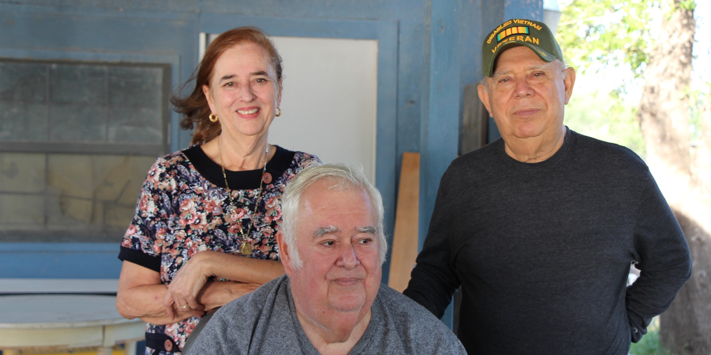 Image: A group of three pose for a photo outside of a wooden house, two standing and one sitting. The woman leans on the chair one man is sitting on. The man standing wears a baseball cap indicating he is a Vietnam War vet.