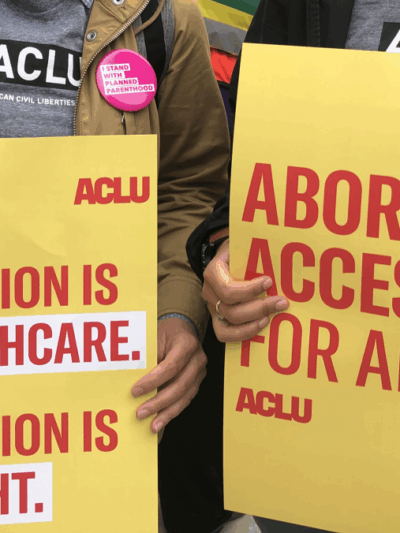 Two people are holding rally signs in support of reproductive freedom. On the left is someone holding a poster that reads "Abotion is healthcare. Aboriton is a right." On the right, a person holds a sign that reads "Abortino Access for All."