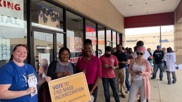 Image: A photo shows people standing in line outdoors along a strip mall. Three women hold a sign that says "Vote to End Mass Incarceration."