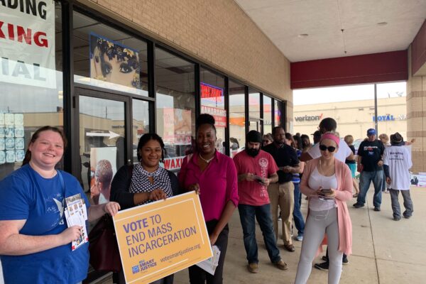 Image: A photo shows people standing in line outdoors along a strip mall. Three women hold a sign that says "Vote to End Mass Incarceration."