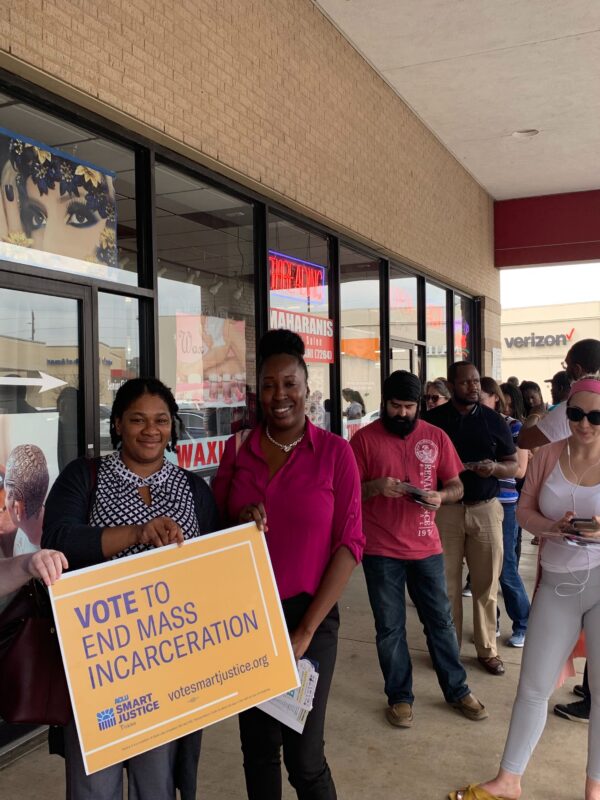 Image: A photo shows people standing in line outdoors along a strip mall. Three women hold a sign that says "Vote to End Mass Incarceration."