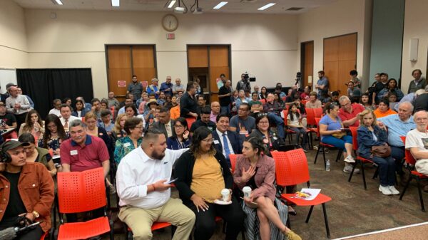 Photo: A crowd of people sit in rows in a large room at the Brownsville Public Library chatting amongst themselves